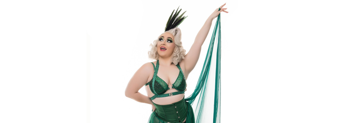 A woman in a green costume and blonde curls hair poses with her hand up in the air